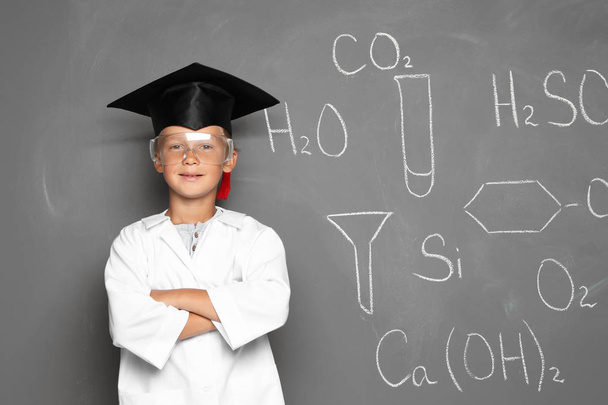 Little school child in laboratory uniform with graduate cap and chemical formulas on grey background - Photo, Image