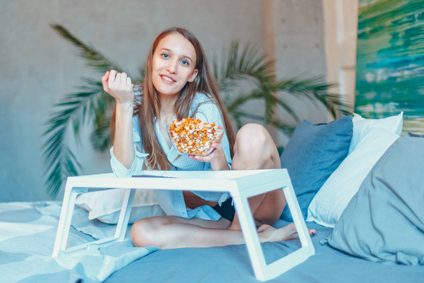 Beautiful young smiling woman watching a movie in the bed and eating popcorn. - Foto, Bild