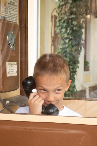 Um rapaz de camisa azul a ligar para o telefone retrô. Rapaz com um telefone antigo na cabine vermelha em estilo inglês. Criança com um auscultador preto. Telefone vintage
. - Foto, Imagem