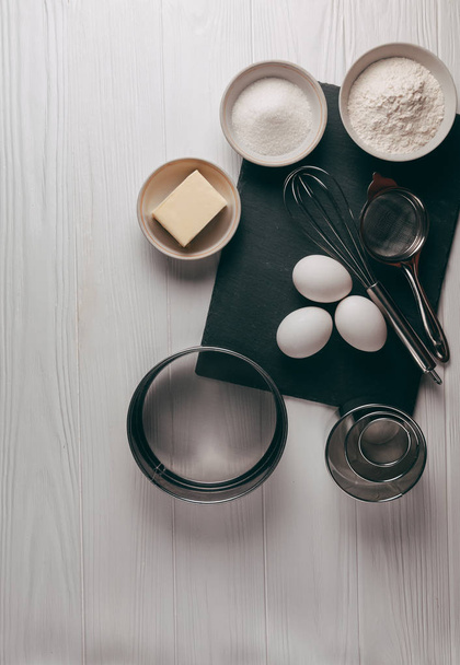 Ingredients and dishes on a white wooden table, which also contains a corolla, are filled with flour, three chicken eggs and a slate board. View from above. Place under the text - Foto, Bild