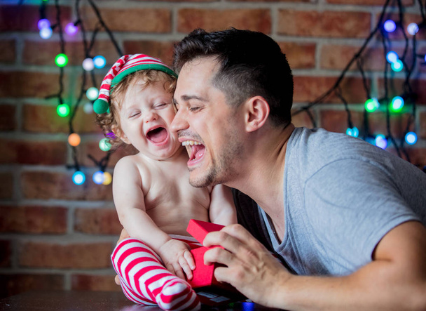 Beautiful little baby boy in elf hat and father, fairy lights on background. Christmas time season image - Photo, Image