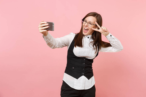 Молодая счастливая деловая женщина в очках делает селфи-снимок на мобильном телефоне, показывая знак победы, выделенный на розовом фоне. Леди-босс. Достижение карьерного богатства. Копирование места для рекламы
 - Фото, изображение