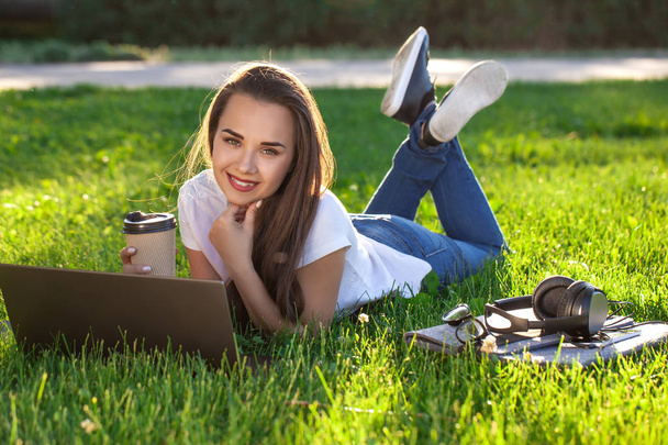 公園でノートパソコンを使っている若い女性が緑の芝生の上に横たわっている。余暇活動のコンセプト. - 写真・画像
