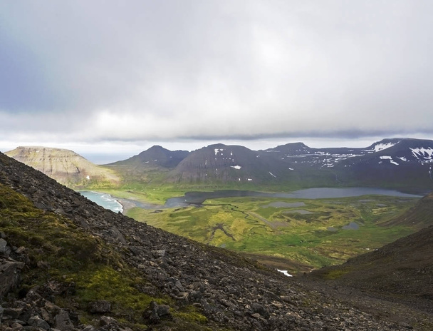 Paisaje de verano del norte, Vista sobre hermosos acantilados cubiertos de nieve y el lago fljotsvatn en la cala de Fljotavik en Hornstrandir, fiordos del oeste, Islandia, con arroyo del río, prado de hierba verde, fondo del cielo malhumorado
 - Foto, Imagen