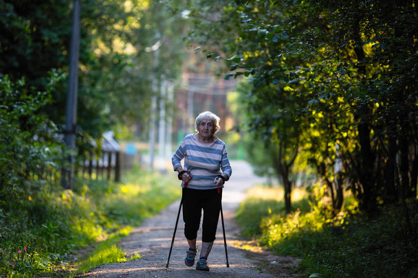 Nordic walking. Elderly woman with ski poles is on the road. (90 years old) - Photo, Image