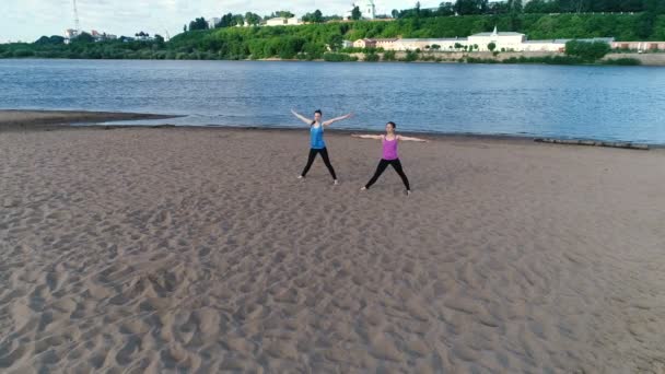 Two woman doing yoga on the sand beach by the river in the city. Beautiful city view in sunrise. - Video, Çekim