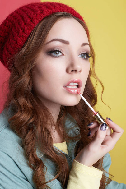fashionable hipster girl in hat, painting lips with lipstick. Beautiful young woman applying makeup. winter fashion. Beauty Model Girl with curly hair and red cap - Photo, image