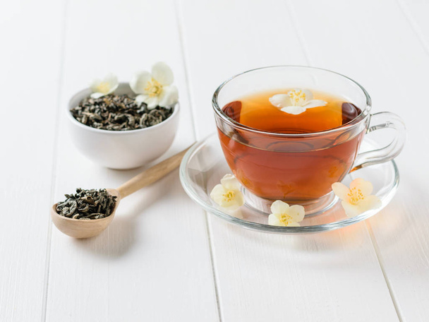 Τσάι από βότανα, μέλι, άνθη γιασεμιού και ένα ξύλινο κουτάλι σε ένα λευκό τραπέζι. Η σύνθεση του το πρωί πρωινό. Άνοιξη τσάι σύνθεση σε έναν λευκό πίνακα. - Φωτογραφία, εικόνα