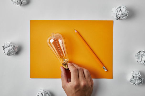白い表面にしわにされたペーパーで囲まれた鉛筆で空白の黄色い紙に白熱ランプを保持している男のショットをトリミング - 写真・画像