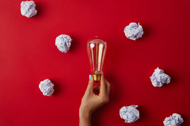 cropped shot of woman holding vintage incandescent lamp surrounded with crumpled papers on red surface - Photo, Image