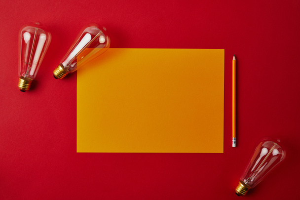 top view of blank yellow paper with pencil and incandescent lamps on red surface - Foto, Imagen