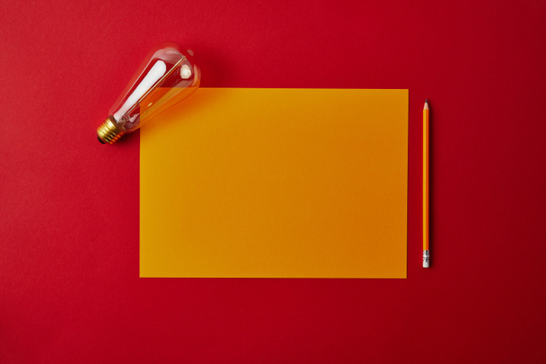 top view of blank yellow paper with pencil and incandescent lamp on red surface - Fotografie, Obrázek