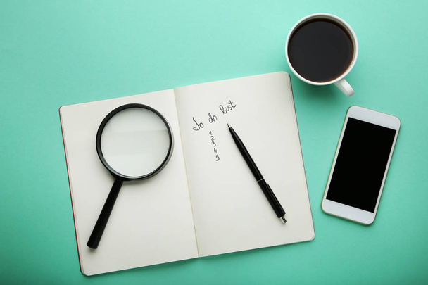 Lista de tareas en bloc de notas con taza de café, teléfono inteligente y lupa sobre fondo de menta
 - Foto, imagen
