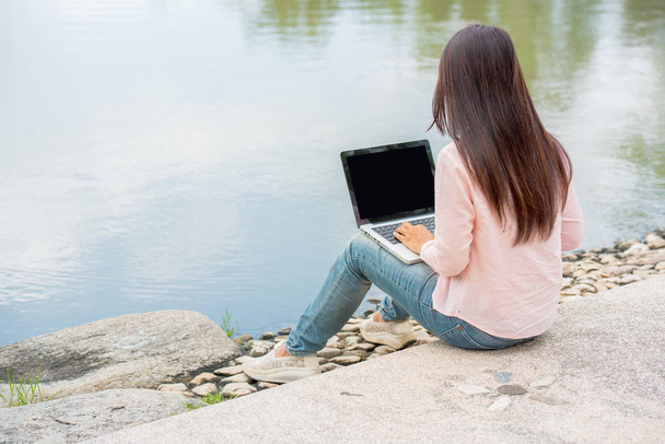 Młoda Azjatka piękna kobieta z uśmiechniętą twarzą pracująca na świeżym powietrzu w parku publicznym. Pracuję na laptopie na zewnątrz. Przycięty obraz kobiety pracującej na laptopie podczas siedzenia w parku. - Zdjęcie, obraz