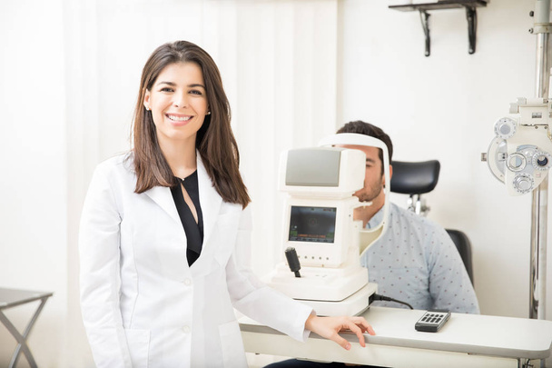 Портрет красивой улыбающейся женщины в халате, стоящей в офтальмологической клинике с мужчиной, сидящим на заднем плане для проверки зрения
 - Фото, изображение