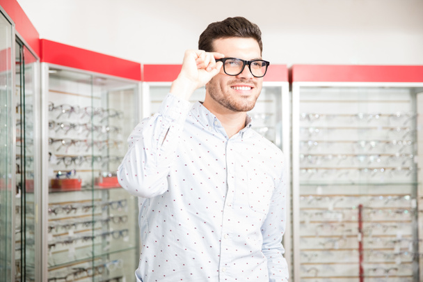 Allegro giovane uomo sorridente mentre tiene la maniglia degli occhiali distogliendo lo sguardo nel negozio ottico con espositore sullo sfondo
 - Foto, immagini