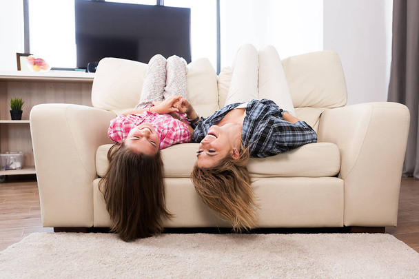 Beautiful mother with her cute daughter lying on the couch with their head upside down - Foto, imagen