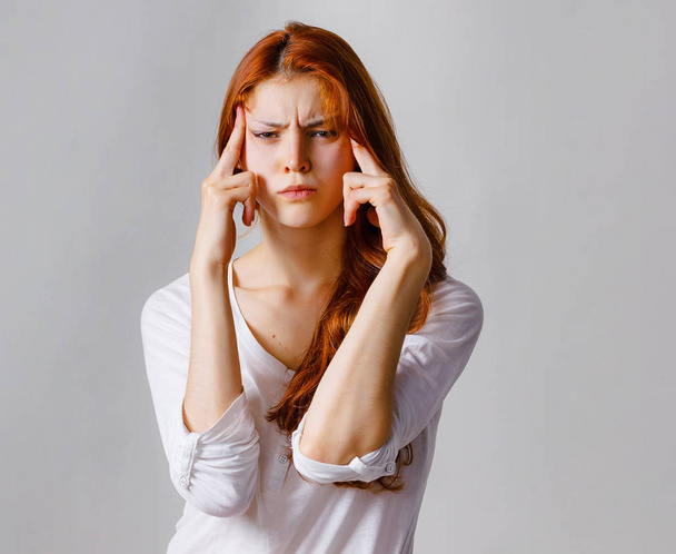 Tender redhead woman has high blood pressure, face expression. Portrait of stressed angry woman holding hands on head. Casual clothes, copy space, gray background - Photo, Image
