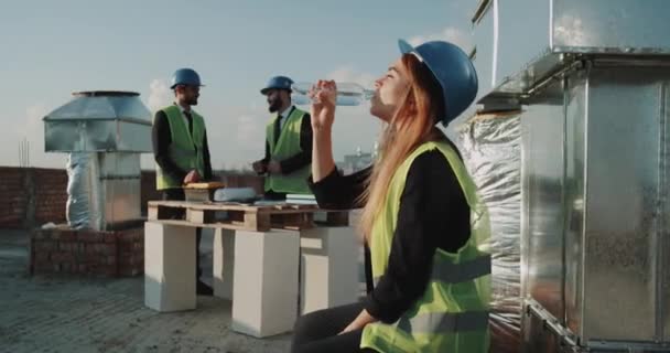 Smiling young woman on construction site , drinking water , wearing a safety helmet and high visibility vest , background builders working - Кадри, відео