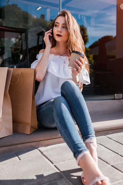 Beautiful stylish young woman with shopping bags having rest on city street and talking on phone. Happy girl drinking coffee by cafe in summer - Photo, Image