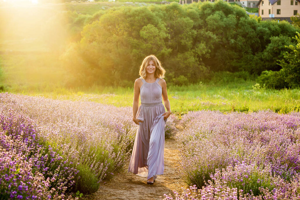 Nuori tyttö violetissa mekossa kävelee laventelipellolla aamuauringossa.
 - Valokuva, kuva