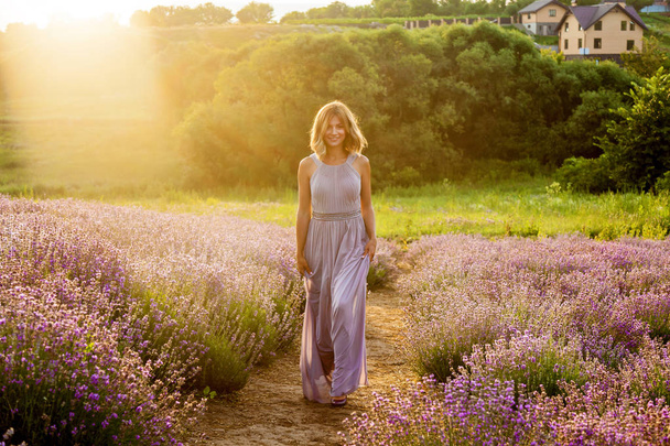 Nuori tyttö violetissa mekossa kävelee laventelipellolla aamuauringossa.
 - Valokuva, kuva
