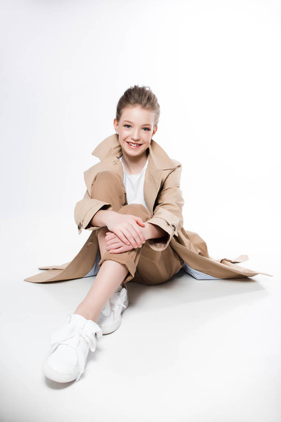 Young girl in beige trench sitting on white background - Zdjęcie, obraz