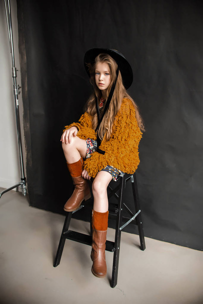 Young girl in orange furry coat and black hat posing on black background in studio - Photo, image