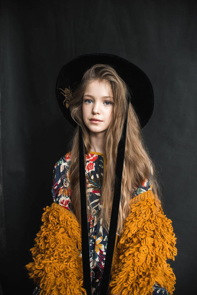 Young girl in orange furry coat and black hat posing on black background in studio - Fotografie, Obrázek