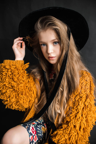 Young girl in orange furry coat and black hat posing on black background in studio - Photo, Image