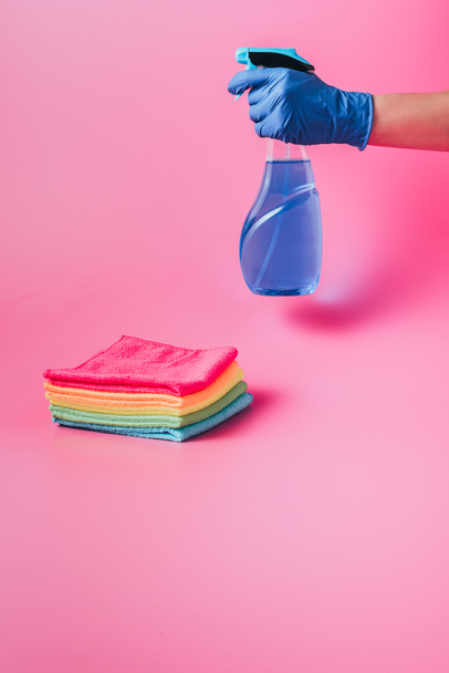 cropped image of female cleaner holding cleaning fluid near stack of colorful rags, pink background - Foto, afbeelding