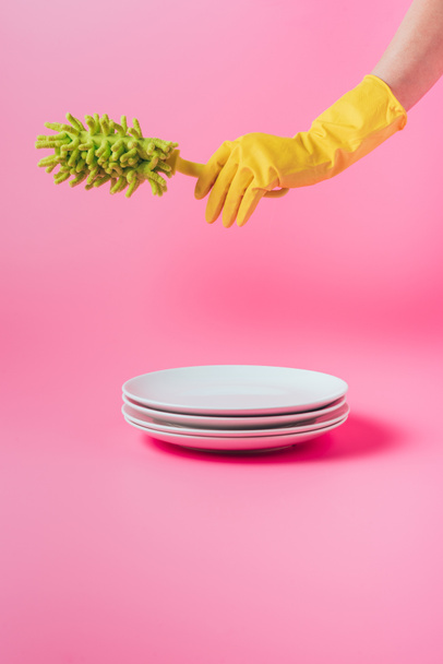 imagen recortada de la mujer en guante de goma celebración plato cepillo de microfibra sobre pila de placas blancas, fondo rosa
  - Foto, Imagen