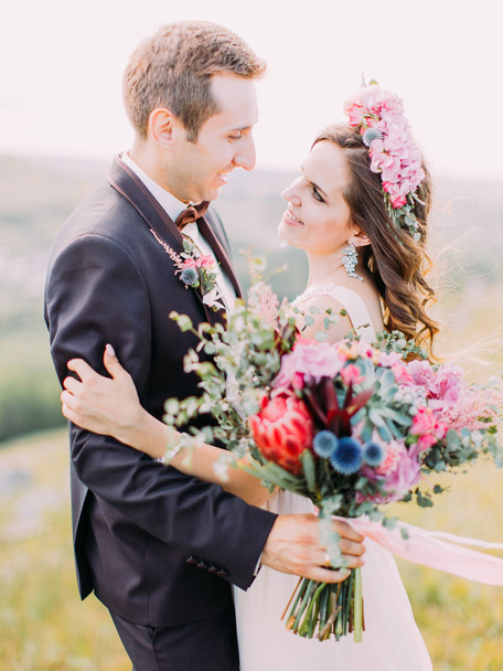Zijaanzicht van de gelukkig knuffelen pasgetrouwden houden de enorme bruiloft boeket van exotische bloemen. - Foto, afbeelding