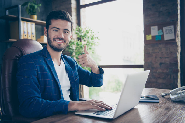 Portrait of attractive handsome young brunet smiling executive worker man, sitting in office workstation workplace, using laptop, showing thumb up - 写真・画像