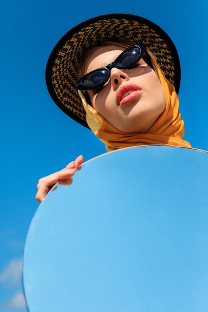 atraktivní stylové dívka pózuje v módní sluneční brýle a drží kulaté zrcadlo s odrazem na modré obloze  - Fotografie, Obrázek