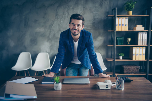 Jovem homem de negócios de casaco azul com barba no rosto fica à mesa no escritório e sorri
 - Foto, Imagem