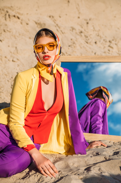 beautiful model in colorful fashionable clothes posing near mirror on sand dune - Foto, Imagem