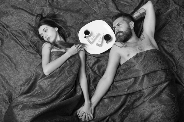 Man with beard and calm face has romantic breakfast with dreamy lady in bed, top view. Man and woman with half covered bodies hold hands - Фото, изображение