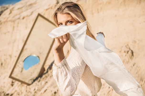 elegáns nő pózol a fehér ruhákat selyemkendőt, közel a tükör a homok - Fotó, kép