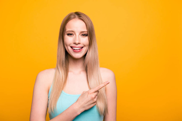 Advertisement concept. Confident young blond woman with beaming smile pointing with her index finger on copy space and looking on camera isolated on shine yellow background - Photo, Image