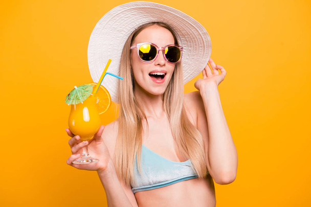 Attractive nice cute straight-haired beautiful blonde caucasian smiling girl, wearing blue swimsuit, sun glasses, hat. Opened mouth, excited, holding cocktail glass. Isolated over yellow background - 写真・画像