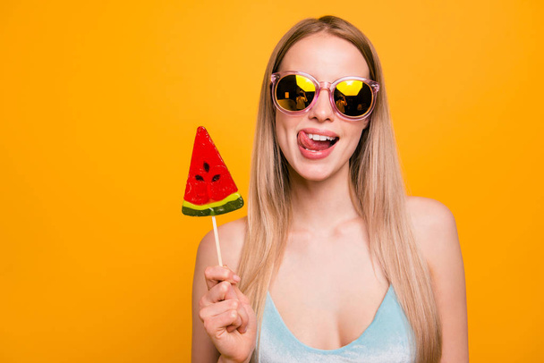Attractive nice cute childish straight-haired beautiful blonde girl, wearing blue swimsuit, color sun glasses, fooling, showing tongue out, holding candy on stick. Isolated over yellow background - Photo, image