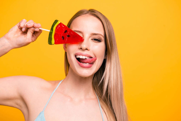 Close up portrait of girl with long blond hair covers her eyes with caramel candy on a stick isolated on shine yellow background - Zdjęcie, obraz