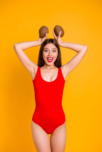 Portrait of charming lady in red swimsuit with coconut in hands above the head isolated on bright yellow background - Foto, afbeelding