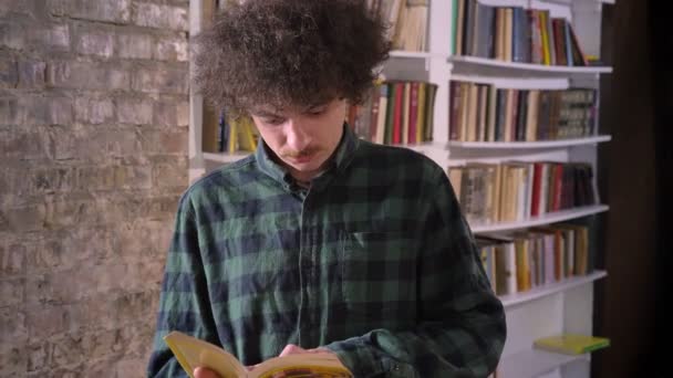 Smart nerdy student with curly hair reading book in library and standing, bookshelves in background - Кадри, відео