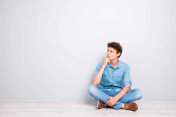 Handsome curly-haired serious young guy wearing casual jeans denim, shoes, sitting on floor with crossed legs, thinking. Copy space. Isolated over light grey background - Foto, imagen