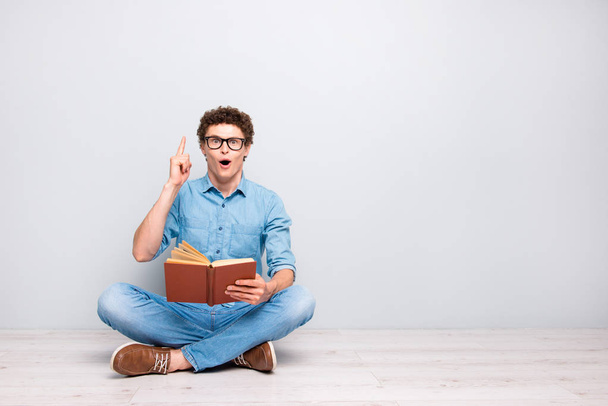 Good thought! Handsome curly-haired amazed guy wearing casual jeans denim, shoes, glasses, sitting on floor with crossed legs, reading book, pointing finger up. Copy space. Isolated on grey background - Foto, Bild