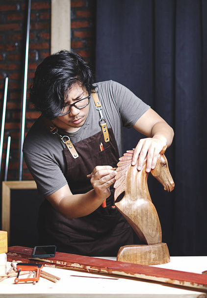 asiatischer Tischler, der in einer Holzbearbeitungswerkstatt arbeitet. Herstellung von hölzernen Pferdeskulpturen - Foto, Bild