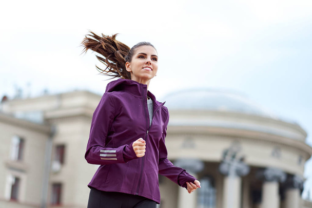 Young female runner in hoody is jogging in city street on background of an architectural building - Foto, Imagem