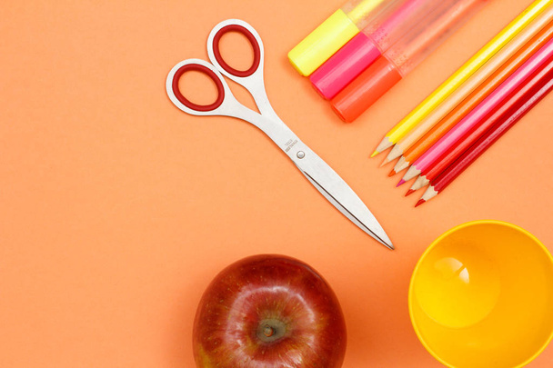 Des fournitures scolaires. Pomme rouge, ciseaux, stylos en feutre, crayons de couleur et tasse en plastique sur fond rose. Vue supérieure avec espace de copie. Retour à l'école. Couleurs pastel
 - Photo, image